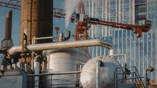 Industrial facility with insulated pipes, a venting stack, and a red tower crane against a glass building