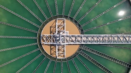Overhead view of a circular clarifier with a central metal bridge and radiating trusses over green water