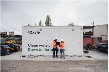 Oxyle branded container unit with two engineers in orange PPE consulting a laptop at an industrial yard
