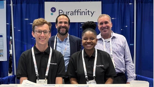 Four people in matching black polo shirts stand smiling behind a trade show booth desk with brochures