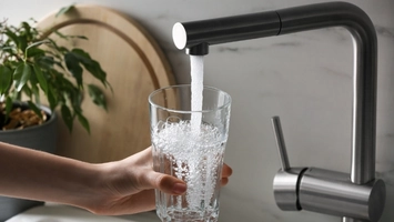 Hand holding glass being filled from modern black kitchen tap with plant and wooden board in background
