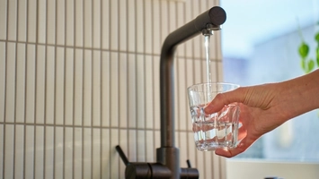A hand holding a glass under a running dark tap against a tiled wall with natural light in the background